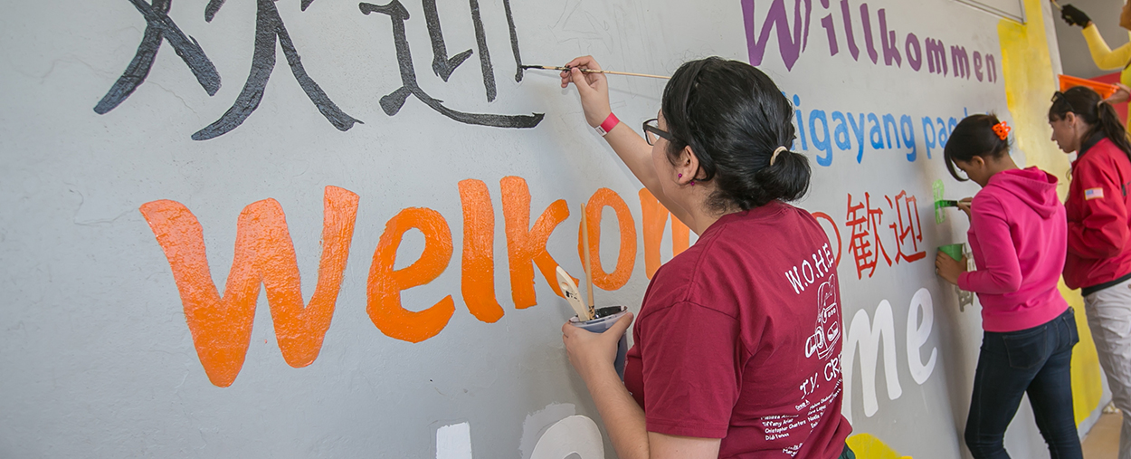 Students painting a wall with Welcome written on it in various languages